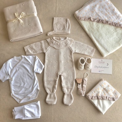 Conjunto de roupa de bebé com peças em malha bege e tecido branco sobre superfície bege