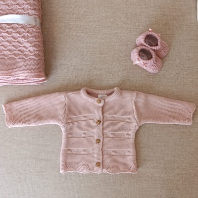 Casaco de malha rosa claro para bebé com botinhas e manta rosa sobre superfície bege