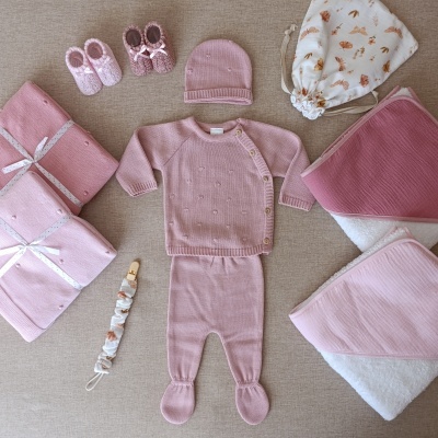 Conjunto rosa claro de roupa e acessórios para bebé sobre superfície bege.