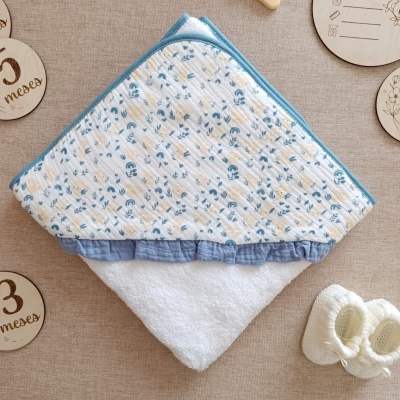 Toalha de bebé com capuz branco e detalhes azuis com sapatilhas brancas e placas de madeira em fundo bege.
