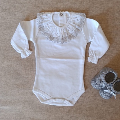 Body branco com gola de renda bordada e sapatos de malha cinza para bebé sobre superfície castanha