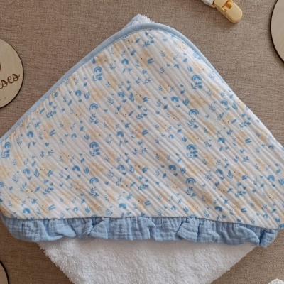 Toalha de bebê com capuz com padrão amarelo e azul, acabamento em tecido azul claro e folho, sobre superfície bege.