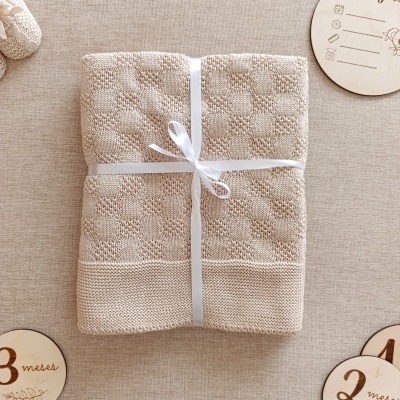 Manta de bebé bege com padrão texturizado e fita branca, com discos de madeira marcando meses