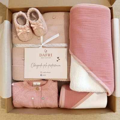 Conjunto de roupa e acessórios de bebé em tons rosa e bege numa caixa de cartão.