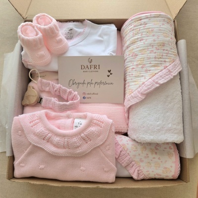 Conjunto de roupa de bebé rosa e branca com botins de malha e manta colorida num recipiente de cartão.