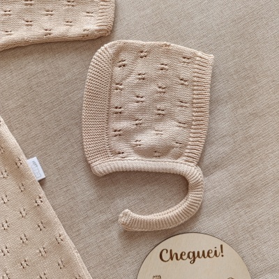 Touca de bebé em malha bege com textura perfurada sobre tecido bege e placa de madeira com texto Cheguei!