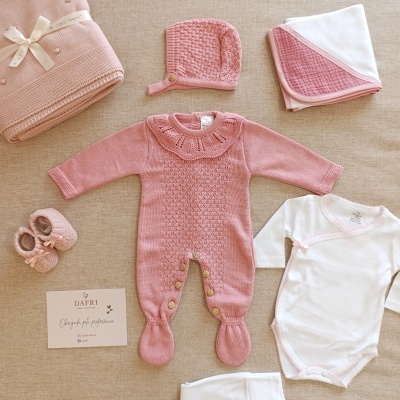Conjunto de roupa e acessórios para bebé em rosa e branco sobre fundo bege.