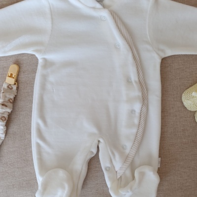 Macacão de bebé branco com botões de pressão, tecido macio, sobre superfície bege com clip de chupeta e brinquedo amarelo