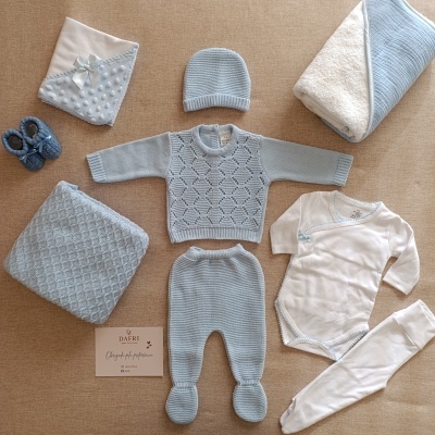 Roupas de bebé em azul claro e branco com texturas tricotadas, mantas e sapatinhos dispostos sobre tecido bege.