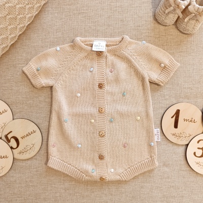 Macacão de bebê bege em malha com botões de madeira e detalhes coloridos, junto a sapatinhos e discos de meses.