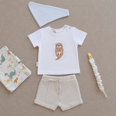 Conjunto roupa bebé com t-shirt branca e calções beges, bandana azul clara, presa de chupeta com padrão e fralda estampada.