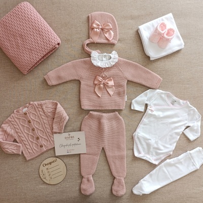 Conjunto de roupa de bebé em rosa e branco com vários acessórios organizados sobre tecido bege.