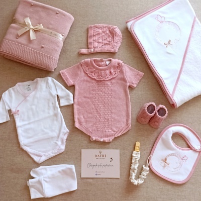 Conjunto de roupa de bebé rosa e branca com diferentes peças e itens em tecido sobre fundo bege.