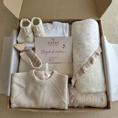 Caixa aberta com roupa e acessórios para bebé em tons bege e branco