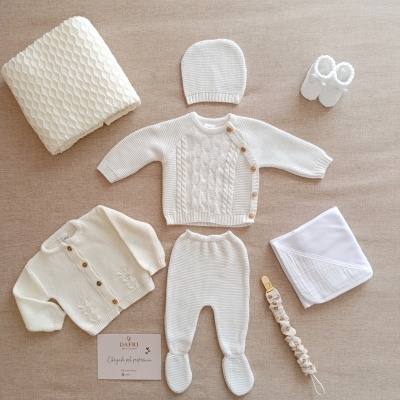 Conjunto de roupa de bebé em malha branca e bege claro com diversos acessórios sobre tecido castanho claro