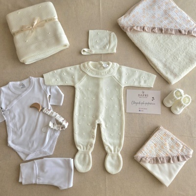 Conjunto de roupa de bebé branco com macacão, body, manta e sapatinhos sobre fundo bege