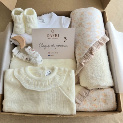 Conjunto de roupa de bebé em caixa com várias peças em tons creme e branco e cartão da marca DAFRI