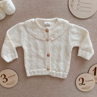 Casaco branco de malha para bebé com botões de madeira sobre tecido bege com placas de madeira
