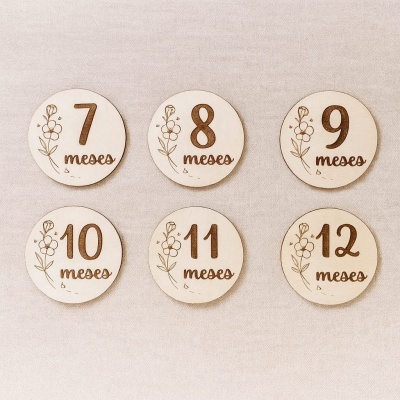Discos redondos de madeira clara com números de 7 a 12 e palavra meses gravados, com desenhos florais, sobre tecido bege.