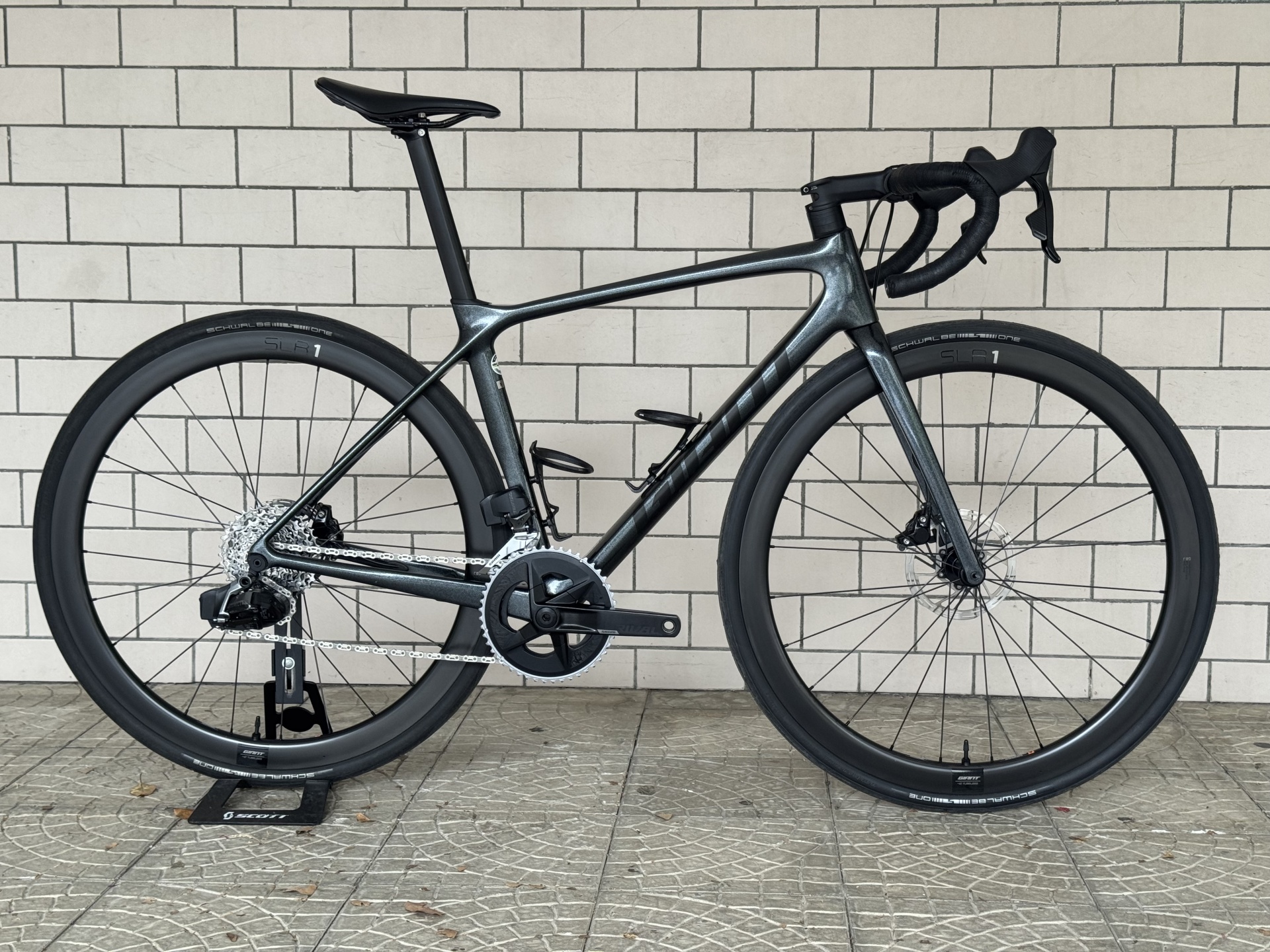 BICICLETA ESTRADA GIANT ADVANCED TCR PRO 1 - RIVAL - USADA Bicicleta de estrada cinza metálica com pneus pretos Schwalbe e azulejos brancos no fundo.
