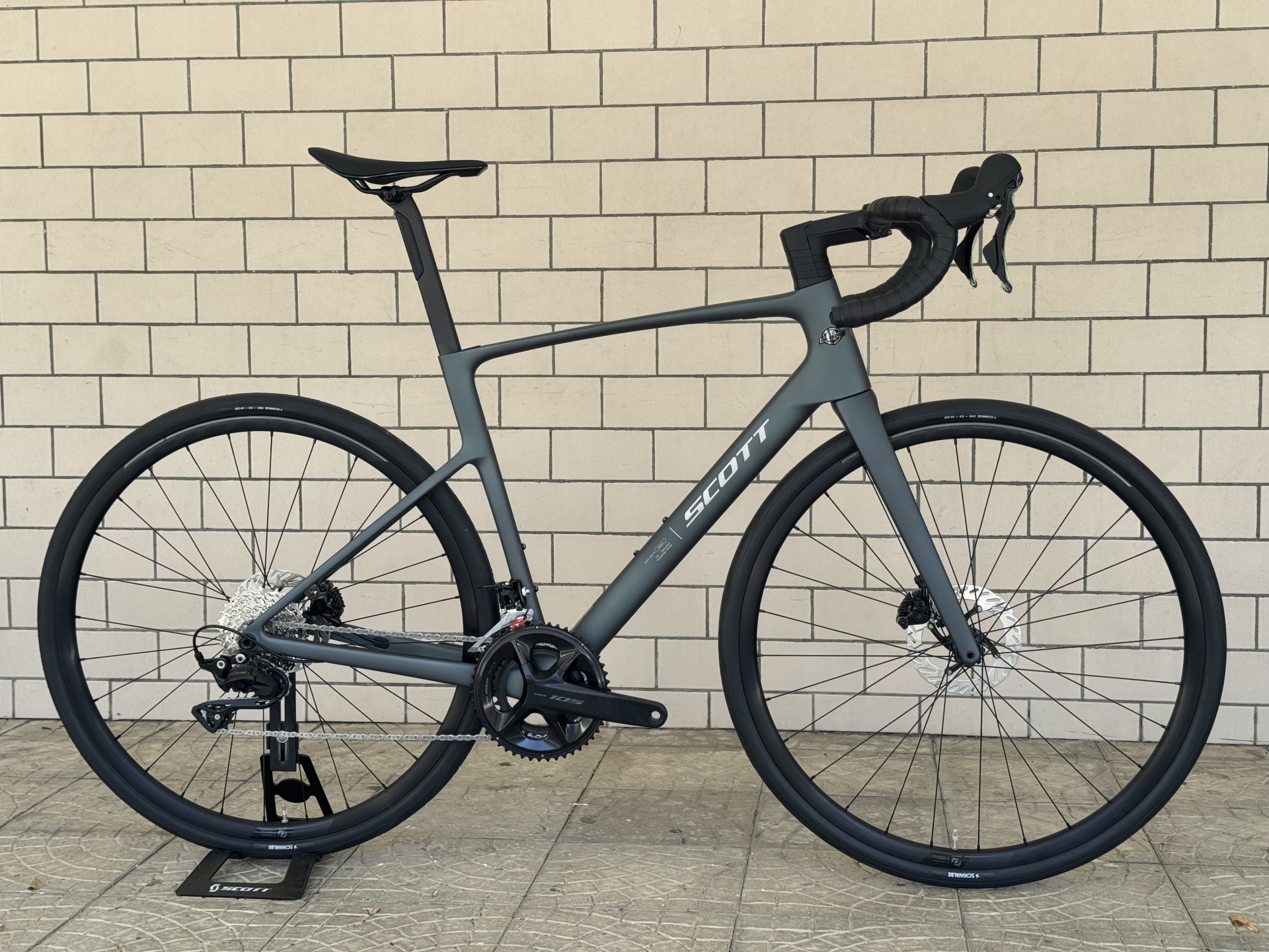 BICICLETA SCOTT ADDICT 50 - CARBON GREY Bicicleta de estrada cinza fosco com detalhes pretos encostada numa parede de azulejos bege