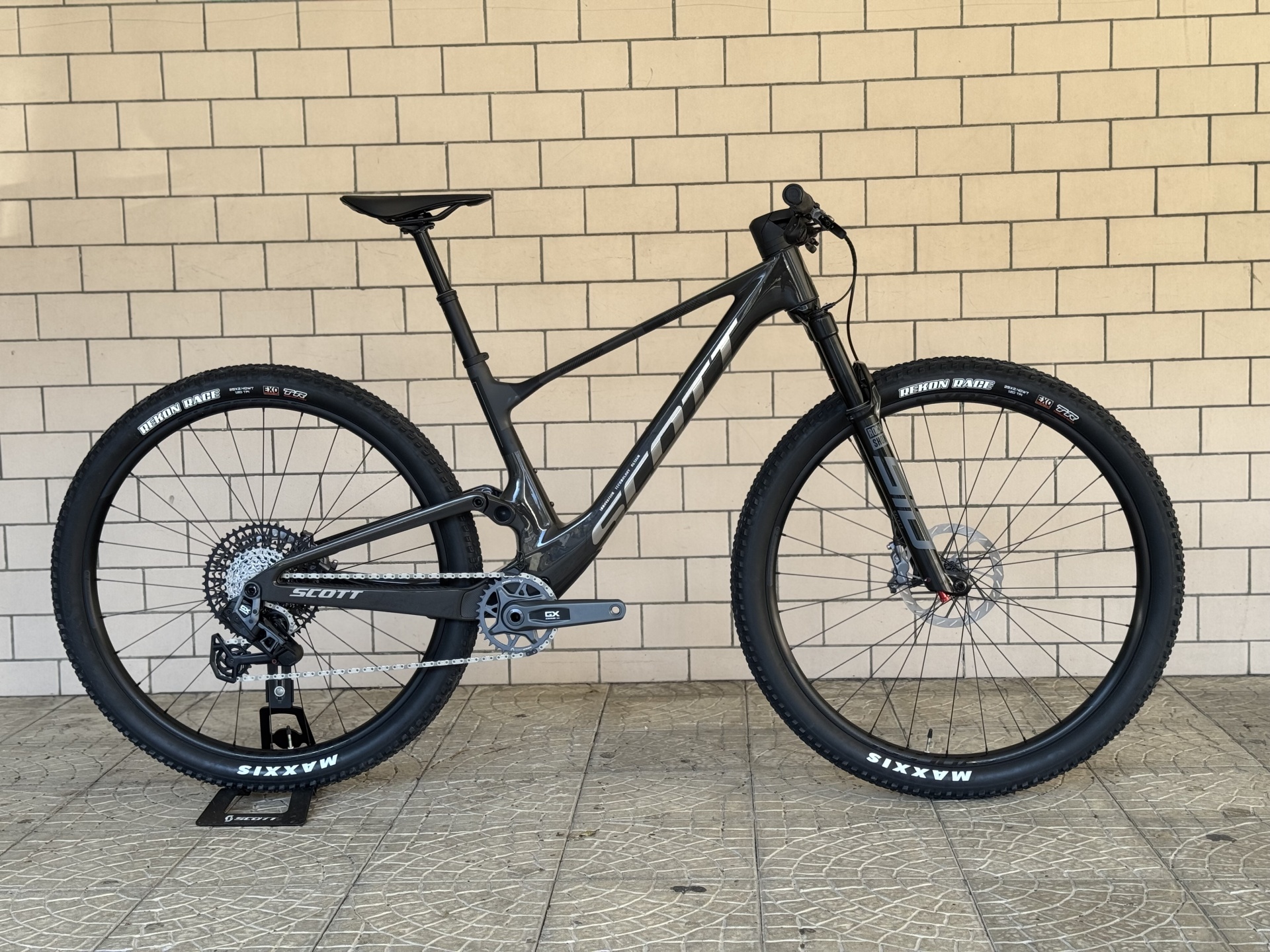 BICICLETA SCOTT SPARK RC TEAM ISSUE - PRETO/CARBONO Bicicleta de montanha preta e cinza com pneus Maxxis junto à parede