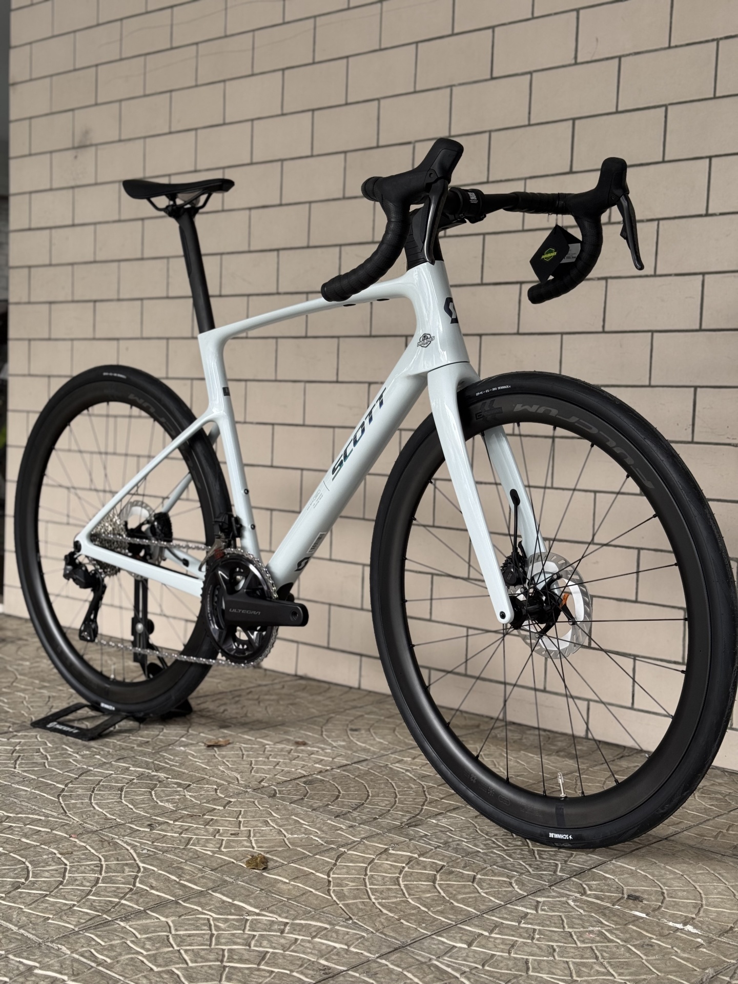 BICICLETA SCOTT ADDICT 20 - CUMULUS WHITE Bicicleta de estrada branca SCOTT com pneus SCHWALBE encostada a parede de azulejos.