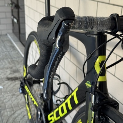 Bicicleta de estrada preta com detalhes amarelos e manetes DURA-ACE ao lado de parede branca revestida a azulejo.