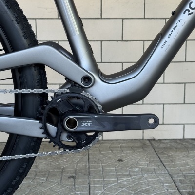 Parte baixa de uma bicicleta de montanha cinza metálica com corrente, roda traseira e pedaleira DEORE XT perto de parede azulejada branca.