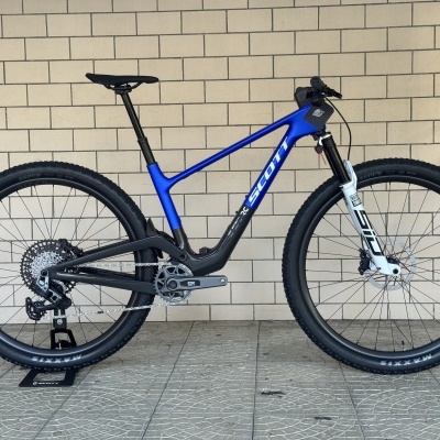 Bicicleta de montanha azul e preta SCOTT com suspensão branca e pneus pretos, apoiada num suporte numa parede de azulejos.
