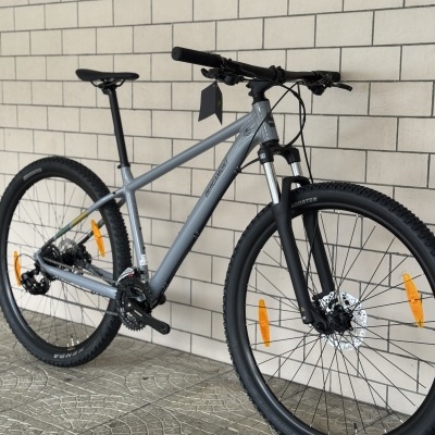 Bicicleta de montanha cinza com suspensão dianteira preta e pneus Kenda junto a uma parede de tijolos claros