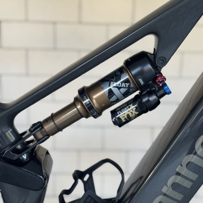 Amortecedor traseiro de bicicleta preto e dourado num quadro Cannondale cinzento escuro junto a uma parede branca em tijolos