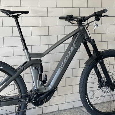 Bicicleta de montanha elétrica cinza com suspensão e pneus Maxxis junto a parede de azulejos brancos