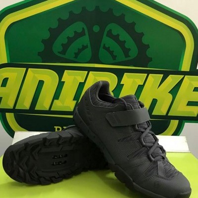 Sapatilhas de ciclismo de montanha pretas com fecho de velcro sobre caixa, em frente a cartaz verde.