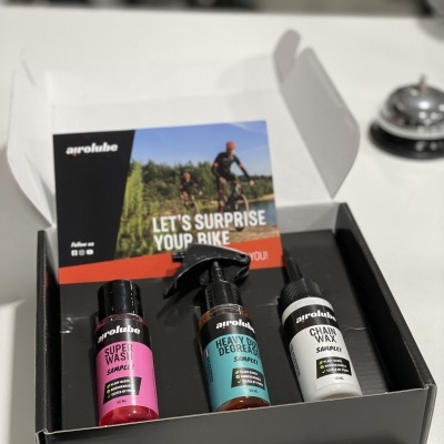 Caixa aberta com três frascos de produtos para bicicleta arolube em cores rosa, azul e branco e folheto promocional