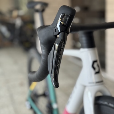 Bicicleta de estrada com punho e cambiador DURA-ACE preto brilhante, quadro branco com detalhes coloridos e roda preta em fundo desfocado.