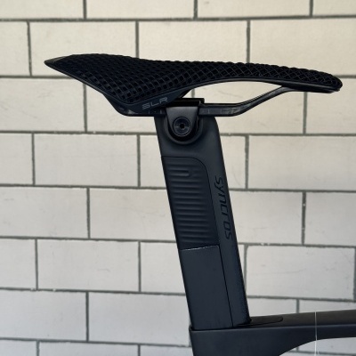 Selim de bicicleta preto texturizado com trem de assento SLR e tubo de selim Syncros preto diante de fundo de azulejos brancos.