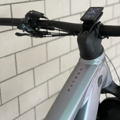Quadro de bicicleta cinza metálico com dispositivo eletrônico no guiador, fundo de azulejos brancos