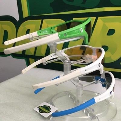 Três óculos de proteção esportiva com armações branca e hastes coloridas em suporte transparente