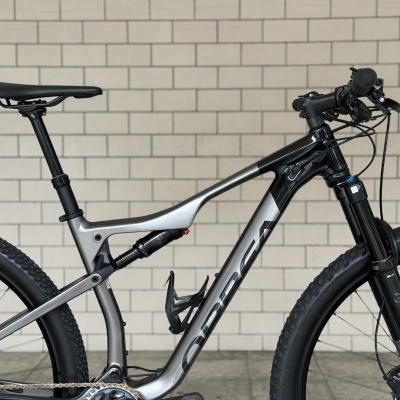 Bicicleta de montanha prateada e preta com pneus pretos e selim preto em frente a parede branca de azulejos