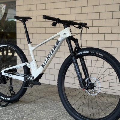 Bicicleta de montanha SCOTT branca com pneus pretos Maxxis e suspensão preta junto a parede de tijolos