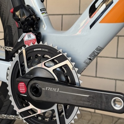 Pedaleira preta com detalhes prateados, janela da corrente de bicicleta branca e laranja com etiquetas vermelhas e escrita SRAM, UCI, RED.