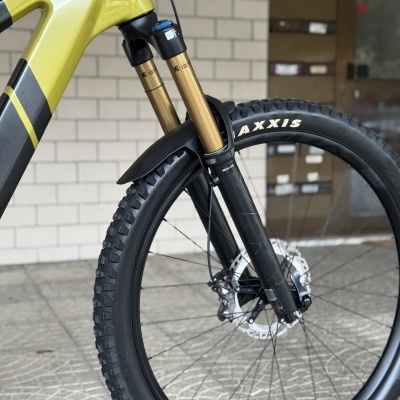 Roda dianteira de bicicleta com suspensão dourada, pneu Maxxis e quadro amarelo e preto, com parede de tijolos ao fundo