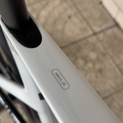 Detalhe do quadro branco de uma bicicleta com texto addict_20, tubo preto e chão azulejado.