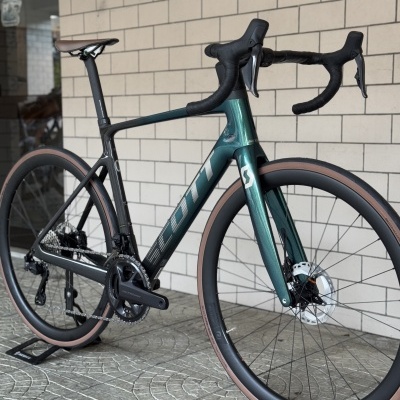 Bicicleta de estrada SCOTT verde e preta com pneus bege à frente de uma parede de tijolos