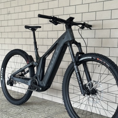 Bicicleta elétrica de montanha cinza escuro e preta apoiada numa parede de tijolos.