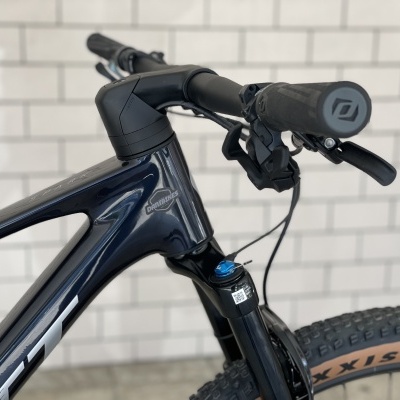 Bicicleta de montanha azul escuro com guiador preto e pneu Maxxis com banda castanha junto a parede azulejos