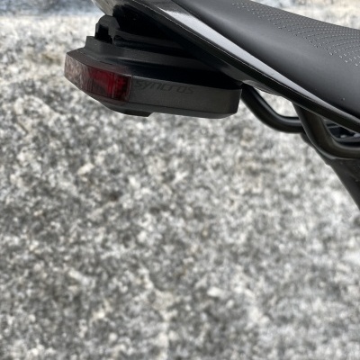 Luz traseira preta com lente vermelha instalada sob banco preto de bicicleta com parede de pedra ao fundo