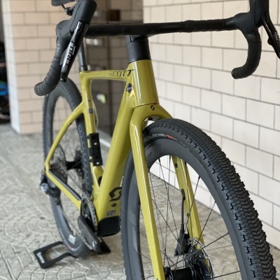 Bicicleta SCOTT verde-oliva com guiador e pneus pretos em suporte, parede de azulejos ao fundo