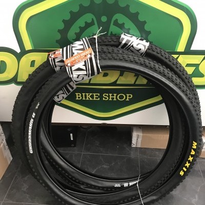 Pneus de bicicleta pretos Maxxis enrolados encostados numa parede com logotipo verde e amarelo